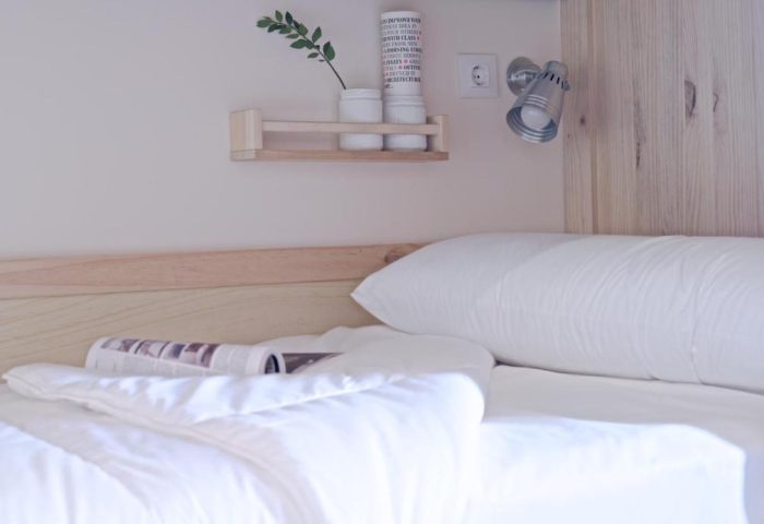 Bedroom detail at The Hat Hostel Madrid, a gay-friendly hotel. Bed with white linens, reading material, shelf with decor, and adjustable reading light.