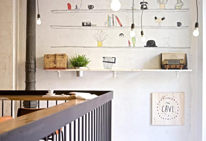 Interior of The Hat Hostel Madrid, a gay-friendly hotel. Shelves with retro decor, drawings, and hanging light bulbs create a stylish, inviting space.