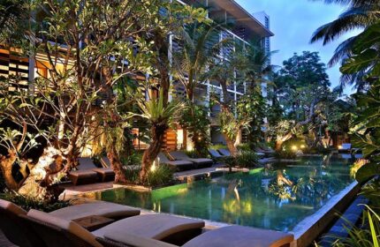 The Haven Bali Seminyak pool at dusk. Gay-friendly hotel with lounge chairs, lush tropical trees, and building in the background. Bali travel.