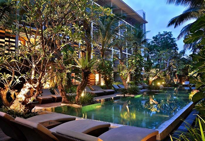 The Haven Bali Seminyak pool at dusk. Gay-friendly hotel with lounge chairs, lush tropical trees, and building in the background. Bali travel.