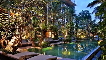 The Haven Bali Seminyak pool at dusk. Gay-friendly hotel with lounge chairs, lush tropical trees, and building in the background. Bali travel.