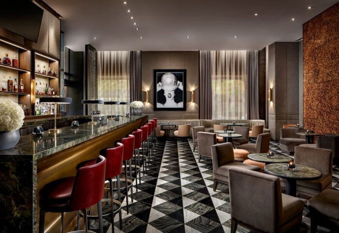 The Hazelton Hotel bar, a gay-friendly spot. Red bar stools line a marble bar. Lounge area with chairs, tables, and patterned floor.