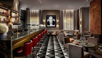The Hazelton Hotel bar, a gay-friendly spot. Red bar stools line a marble bar. Lounge area with chairs, tables, and patterned floor.