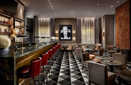 The Hazelton Hotel bar, a gay-friendly spot. Red bar stools line a marble bar. Lounge area with chairs, tables, and patterned floor.