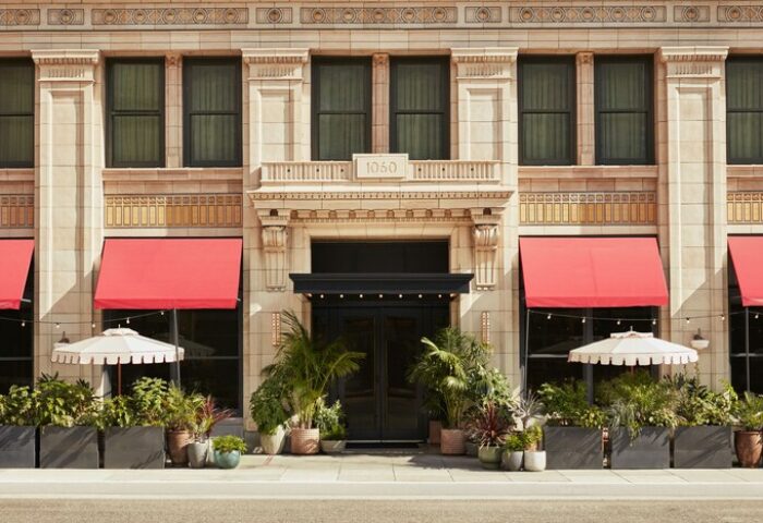 Facade of The Hoxton Downtown LA, a gay-friendly hotel. Red awnings, potted plants, and umbrellas adorn the entrance. Travel advice for gay men.