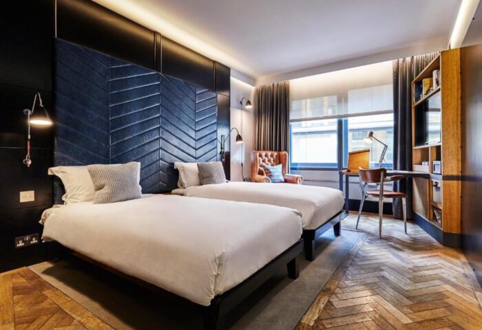 Gay-friendly hotel room at The Hoxton, Shoreditch. Two beds with white linens, herringbone headboard, desk, and chair. Stylish interior design.