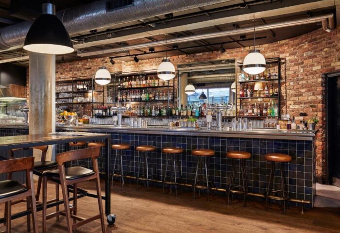 Bar area at The Hoxton, Shoreditch. Gay-friendly hotel with stools lined up at the bar. Shelves stocked with bottles and glasses. Brick wall.