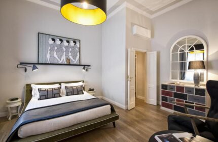 The Independent Suites gay-friendly hotel room. A bed with gray pillows and a gray blanket, a mirror, and a black and yellow lamp.