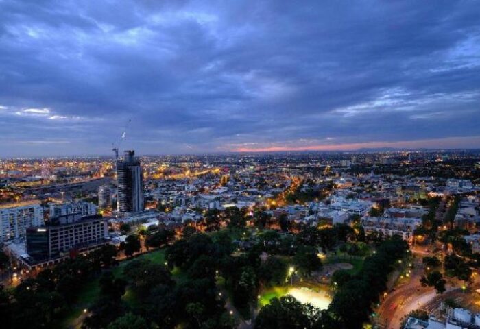 Night view of The Jazz Corner Hotel, a gay-friendly hotel. City lights sparkle under a dramatic sky. Park in foreground. Melbourne skyline.