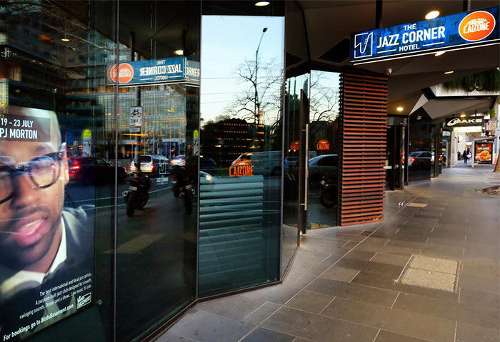 The Jazz Corner Hotel entrance, a gay-friendly hotel. Its glass reflects the street. A poster of PJ Morton is visible. Calzone restaurant nearby.
