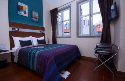 The Keong Saik Hotel room. Gay-friendly hotel bedroom with bed, colorful blanket, two windows, and suitcase. Gay travel in Singapore.