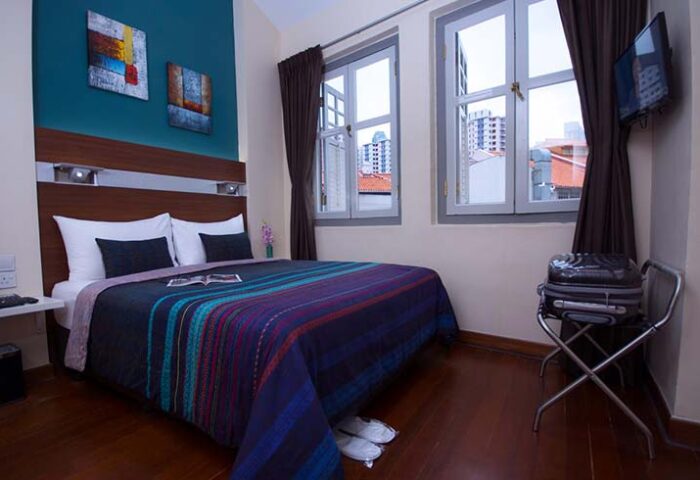 The Keong Saik Hotel room. Gay-friendly hotel bedroom with bed, colorful blanket, two windows, and suitcase. Gay travel in Singapore.