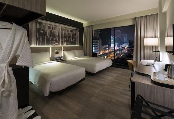 The Kuala Lumpur Journal Hotel room. Gay-friendly hotel with two beds, a desk, and a city view at night. Robe and slippers visible.
