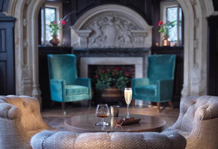 Lalit London gay-friendly hotel lounge. Teal chairs flank a fireplace. Drinks on a table, cozy seating. Luxury gay travel in London.