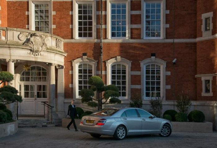 The Lalit London, gay-friendly hotel. Exterior view of the luxury hotel, featuring a car, entrance, and a staff member. Brick building.