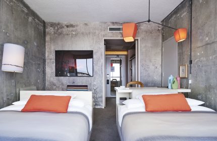 The Linq Hotel and Casino room with two beds, concrete walls, and orange accents. A gay-friendly hotel option in Las Vegas.