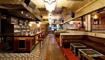 Interior of The Linq Hotel and Casino bar, a gay-friendly hotel. Long bar with stools, leather couches, and stage lighting.