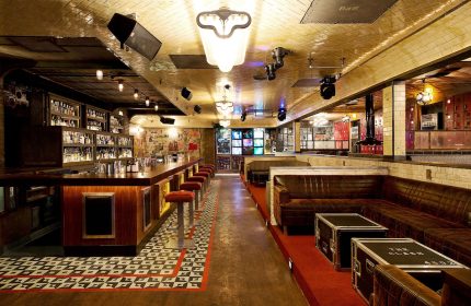 Interior of The Linq Hotel and Casino bar, a gay-friendly hotel. Long bar with stools, leather couches, and stage lighting.