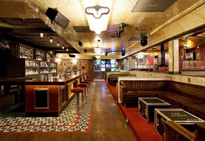 Interior of The Linq Hotel and Casino bar, a gay-friendly hotel. Long bar with stools, leather couches, and stage lighting.