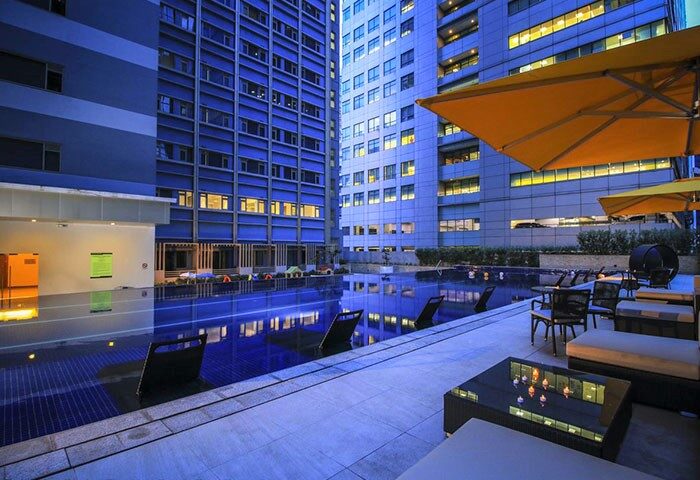 Pool area at The Mini Suites Eton Tower Makati, a gay-friendly hotel. Modern building backdrop, sun umbrellas, & lounge seating.