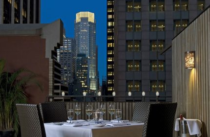 The Muse New York, a gay-friendly hotel. Rooftop dining with city views. Table set with wine glasses, skyscrapers at dusk.