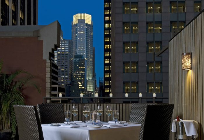 The Muse New York, a gay-friendly hotel. Rooftop dining with city views. Table set with wine glasses, skyscrapers at dusk.