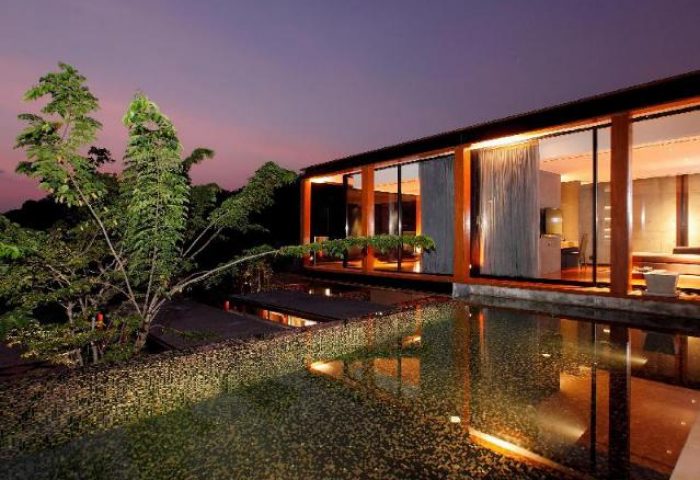 The Naka Phuket Villa, a gay-friendly hotel. Modern villa with a pool reflecting the interior lights at dusk. Tropical trees nearby.