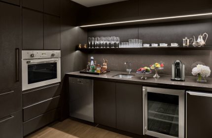 Kitchenette at The New York EDITION, a gay-friendly hotel. Dark cabinets, oven, sink, wine fridge, and shelf with glassware. Upscale amenities.