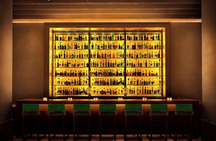 Bar at The New York EDITION, a gay-friendly hotel. Backlit shelves display liquor bottles. Green stools line the bar. Nightlife for gay men.
