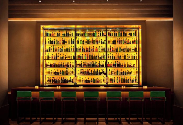 Bar at The New York EDITION, a gay-friendly hotel. Backlit shelves display liquor bottles. Green stools line the bar. Nightlife for gay men.