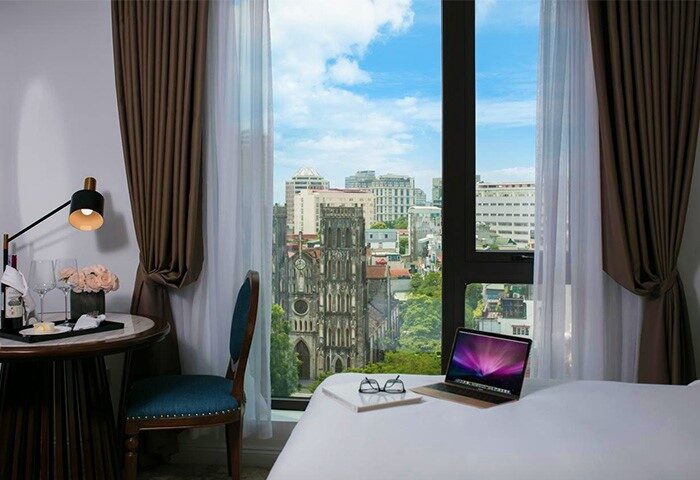 The Oriental Jade Hotel gay-friendly room with city view. Laptop, glasses, and notepad on bed. Hanoi Cathedral visible. Gay travel.