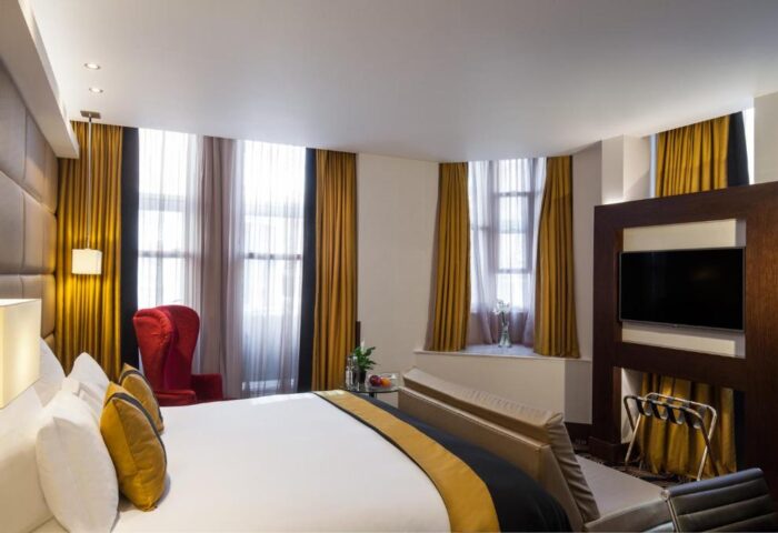 Gay-friendly hotel The Piccadilly London West End: A stylish room with a large bed, red chair, TV, and bright windows. Ideal for gay travelers.