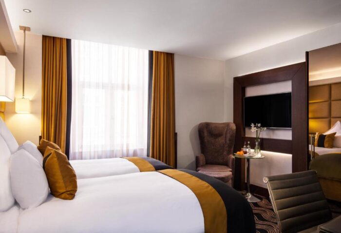 The Piccadilly London West End gay-friendly hotel room. Two beds with gold accents, TV, and chair. Upscale lodging in London's West End.