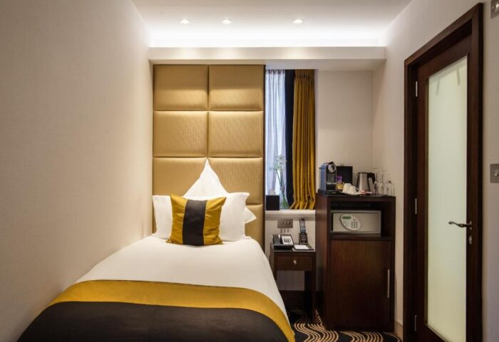 The Piccadilly London West End gay-friendly hotel room. Single bed with gold accents, safe, coffee maker, and a window with curtains.