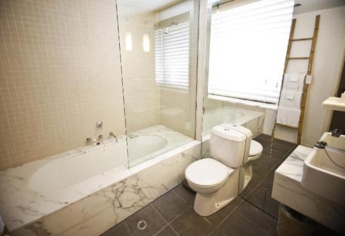 Bathroom at The Prince Hotel, a gay-friendly hotel. Bathtub with glass partition, toilet, sink, and ladder with towels. Modern design.