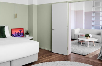 The Prince Hotel gay-friendly room: bedroom with bed, nightstand, and art, leading to a living room with couch, table, and shelves. Modern design.