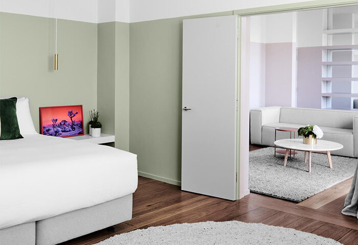 The Prince Hotel gay-friendly room: bedroom with bed, nightstand, and art, leading to a living room with couch, table, and shelves. Modern design.