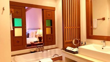 The Rim Chiang Mai Hotel bathroom. Gay-friendly hotel with bathtub, sink, and wooden details. Bedroom view through colorful window.