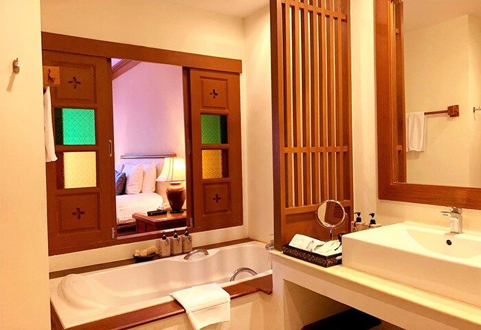 The Rim Chiang Mai Hotel bathroom. Gay-friendly hotel with bathtub, sink, and wooden details. Bedroom view through colorful window.