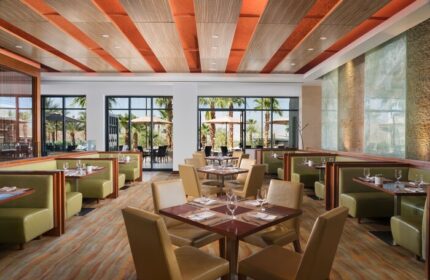 Dining area at The Ritz-Carlton, Rancho Mirage, a gay-friendly hotel. Tables are set, booths line the walls, and windows offer views of palm trees.