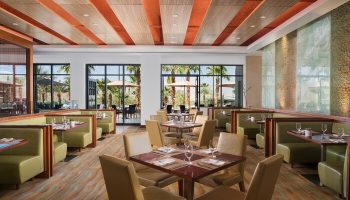 Dining area at The Ritz-Carlton, Rancho Mirage, a gay-friendly hotel. Tables are set, booths line the walls, and windows offer views of palm trees.
