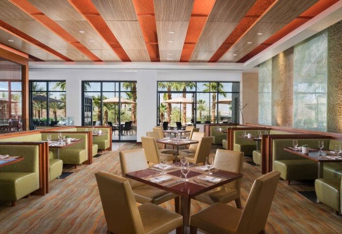 Dining area at The Ritz-Carlton, Rancho Mirage, a gay-friendly hotel. Tables are set, booths line the walls, and windows offer views of palm trees.