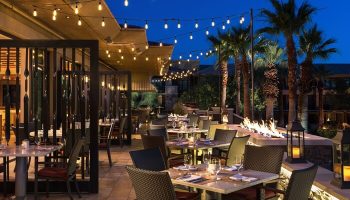 Outdoor dining at The Ritz-Carlton, Rancho Mirage, a gay-friendly hotel. Tables set, fire pit, palm trees, and string lights create ambiance.
