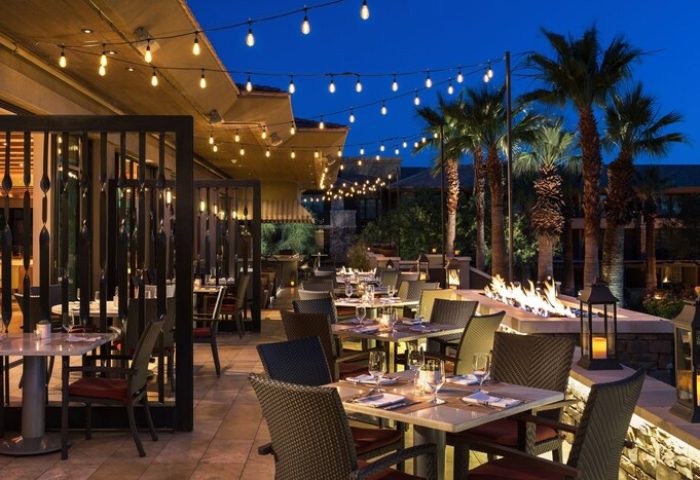 Outdoor dining at The Ritz-Carlton, Rancho Mirage, a gay-friendly hotel. Tables set, fire pit, palm trees, and string lights create ambiance.