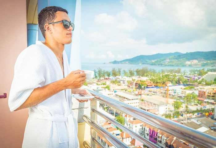 Man in robe at The Royal Paradise Hotel & Spa, a gay-friendly hotel. He's on a balcony, enjoying the view with a drink. Phuket gay travel.