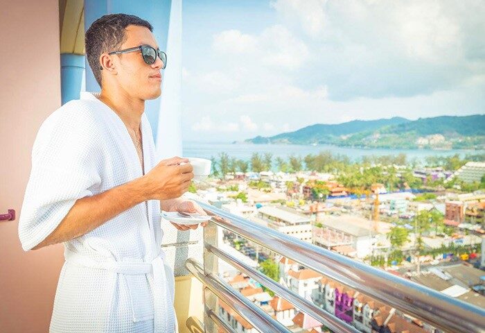 Man in robe at The Royal Paradise Hotel & Spa, a gay-friendly hotel. He's on a balcony, enjoying the view with a drink. Phuket gay travel.