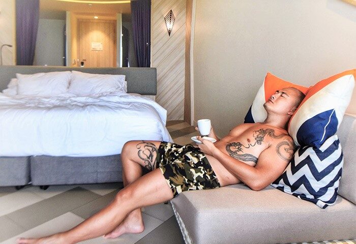 Gay-friendly The SIS Kata Resort: Man relaxes with coffee in a stylish room. Tattoos, modern decor, and a comfortable vibe. Phuket travel.