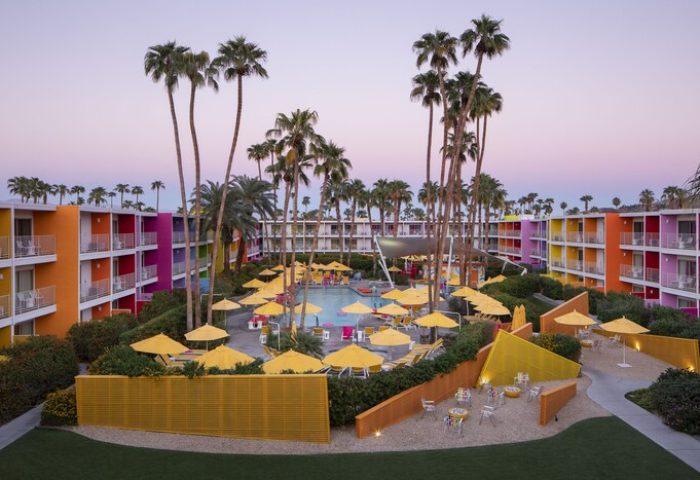 The Saguaro Palm Springs gay-friendly hotel view. Colorful building, pool with yellow umbrellas, and palm trees. A great gay travel destination.