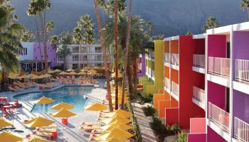 The Saguaro Palm Springs, a gay-friendly hotel. Colorful hotel exterior, pool, yellow umbrellas, and palm trees against a mountain backdrop.