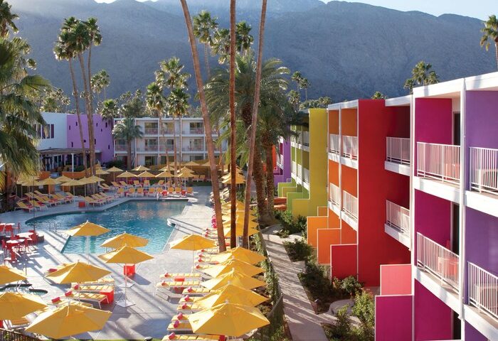 The Saguaro Palm Springs, a gay-friendly hotel. Colorful hotel exterior, pool, yellow umbrellas, and palm trees against a mountain backdrop.